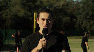 Tom EGE, micro en main, présente le reportage face caméra sur le terrain d'entraînement des Comanches ; des joueuses en tenue noire s'échauffent derrière lui.